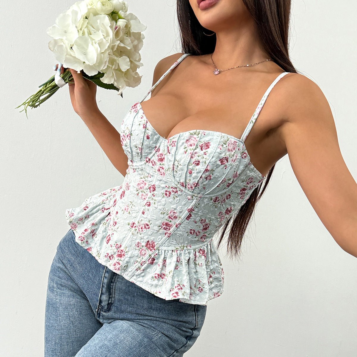 Alt text:
A woman wearing a yellow floral corset-style top with ruffled hem and thin straps, paired with white pants, holding a bouquet of white flowers against a neutral background.