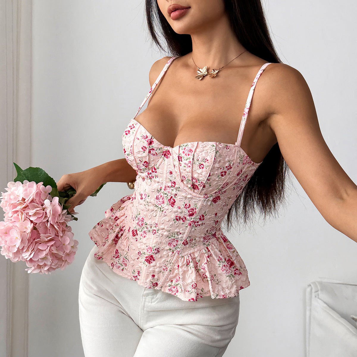 Alt text:
A woman wearing a yellow floral corset-style top with ruffled hem and thin straps, paired with white pants, holding a bouquet of white flowers against a neutral background.